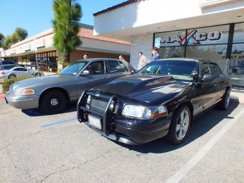 2003 Ford Crown Victoria - Black modified Crown Victoria beside a silver one in a strip-mall lot fronting the Maxco shop, people standing nearby