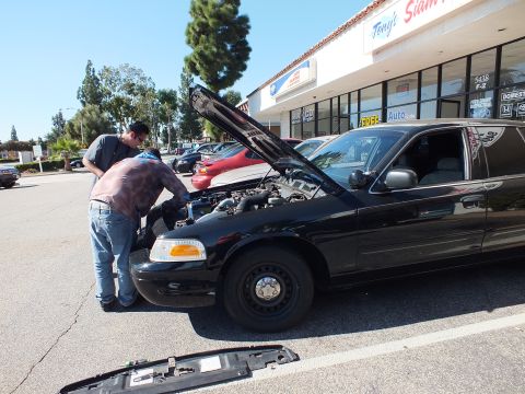 2003 Ford Crown Victoria - Mechanic working under the open hood of a black Crown Victoria, steel wheels exposed, bumper cover on the ground