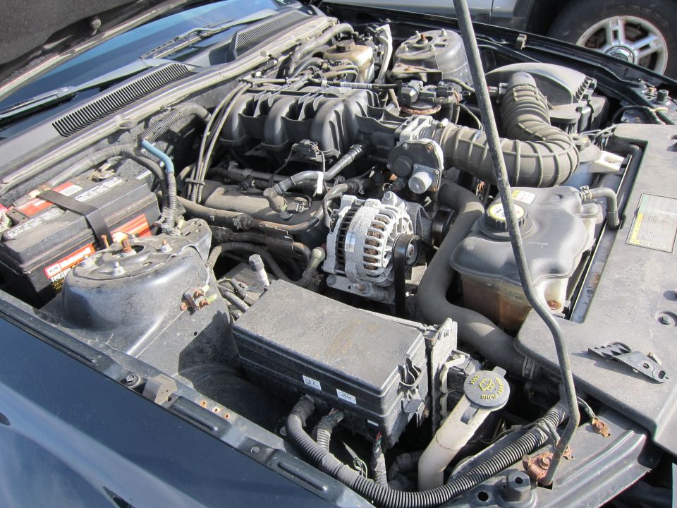 Engine bay with battery, alternator, and various hoses and wiring. A fuse box is visible