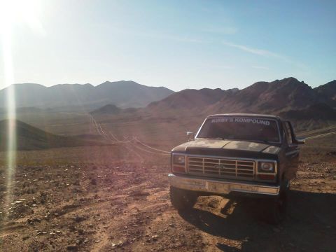 1985 Ford Bronco - A dark Ford truck with a tan hood sits on a dirt road, with mountains in the background under a bright sky.