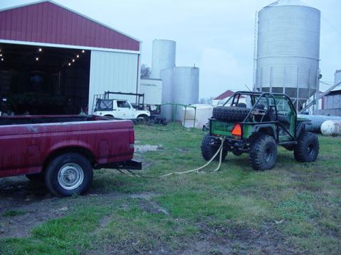 1992 Ford Bronco - A green off-road vehicle with a roll cage and spare tire is being towed by a red pickup truck in a rural setting with silos and a barn.
