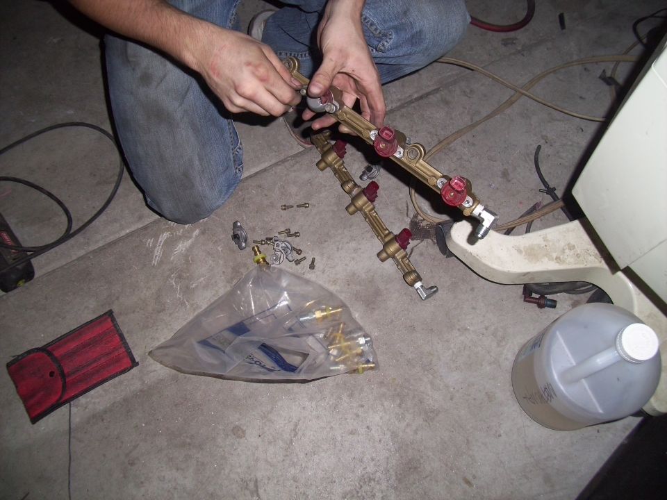 Hands assembling a brass fuel rail with red-capped injectors and fittings. Bag of parts and a plastic jug are nearby