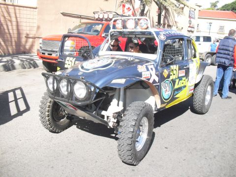 1986 Ford Bronco - A custom off-road buggy with numerous sponsor decals is parked on a street, with a red pickup truck behind it.