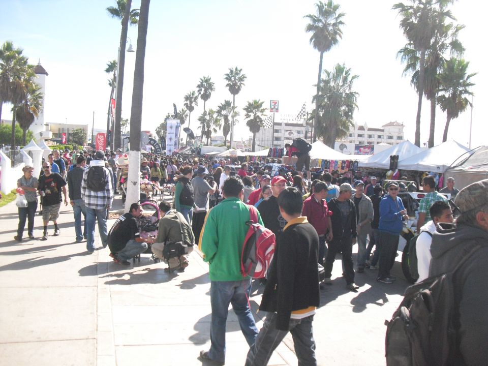 Crowded outdoor event with palm trees and tents, people walking on a sidewalk
