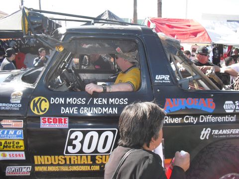 1986 Ford Bronco - A black off-road truck with numerous sponsor decals and racing numbers is seen from the side with drivers inside.
