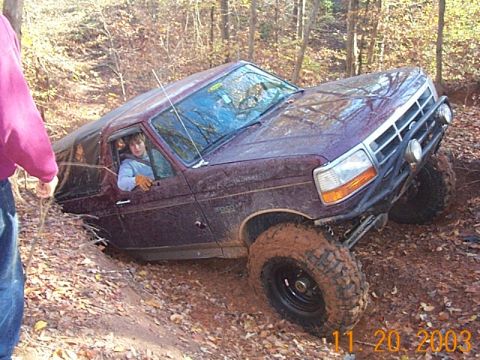1986 Ford Bronco - A maroon Ford Bronco is stuck in a muddy ditch with trees in the background.