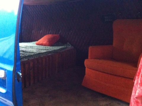 1977 Ford E350 - The interior of a van conversion features a bed with a red pillow and an orange armchair.