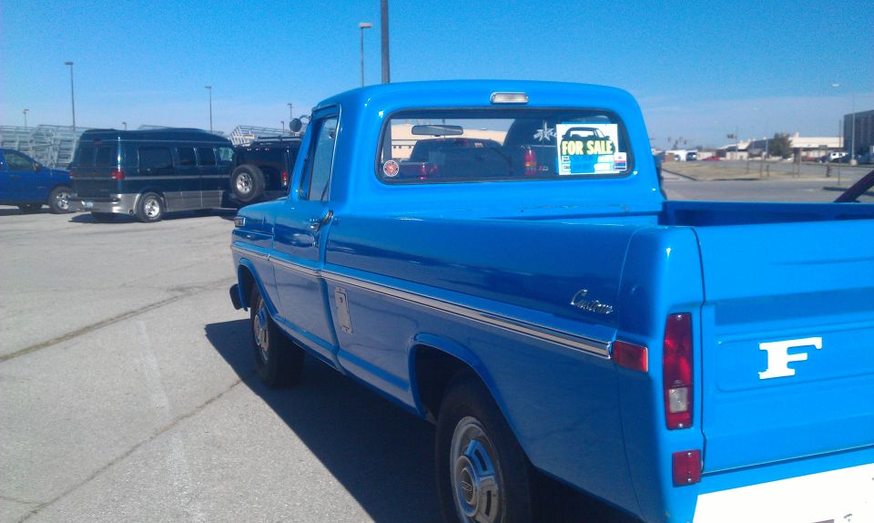 1973 Ford F-100 pickup truck, blue, with a "For Sale" sticker on the rear window. Other vehicles are parked nearby