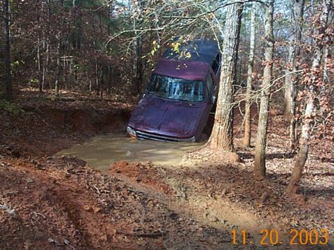 1986 Ford Bronco - A purple SUV is stuck in a muddy hole off-road, with trees and foliage all around.