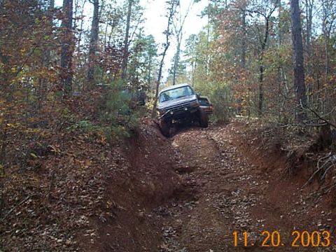 1986 Ford Bronco - A 1986 Ford Bronco navigates a steep, muddy trail in a wooded area, with another vehicle following closely following.