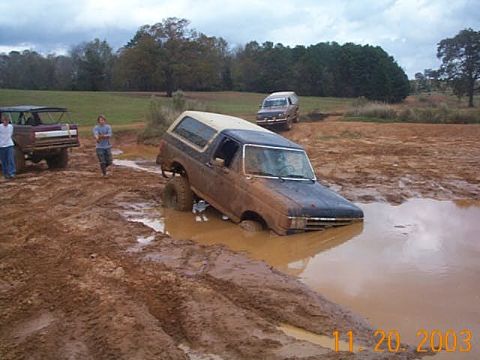 1986 Ford Bronco - Two SUVs navigate a muddy off-road course, one deeply stuck in a water hazard.