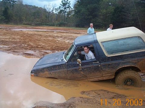 1986 Ford Bronco - A Ford Bronco is submerged in mud, with two people standing nearby watching.