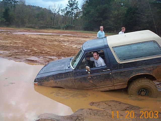 Blue SUV with tan hardtop stuck nose-down in a red clay mud pit, driver leaning out window