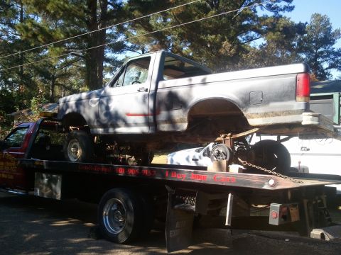 1990 Ford Bronco - A tow truck transports a weathered Ford F-Series pickup truck, likely from the early 1990s.