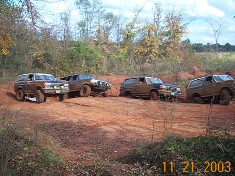 1986 Ford Bronco - Multiple Ford Bronco SUVs navigate a muddy off-road course during daylight hours.