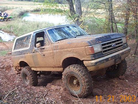 1986 Ford Bronco - A muddy 1986 Ford Bronco navigates a challenging off-road course with other ATVs visible in the background.