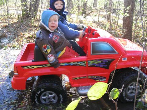 1986 Ford Bronco - Children play in a red toy truck with flame graphics, navigating a wooded area with water.