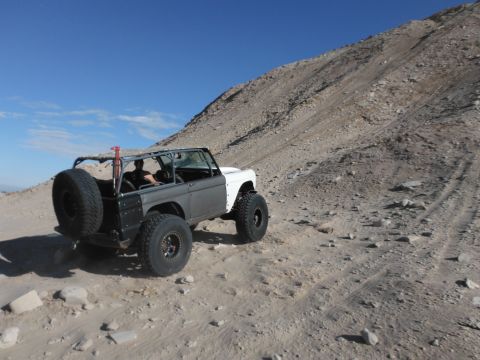 1979 Ford Bronco - Off-road Bronco with custom roll cage navigates a steep, rocky desert trail under a clear blue sky.