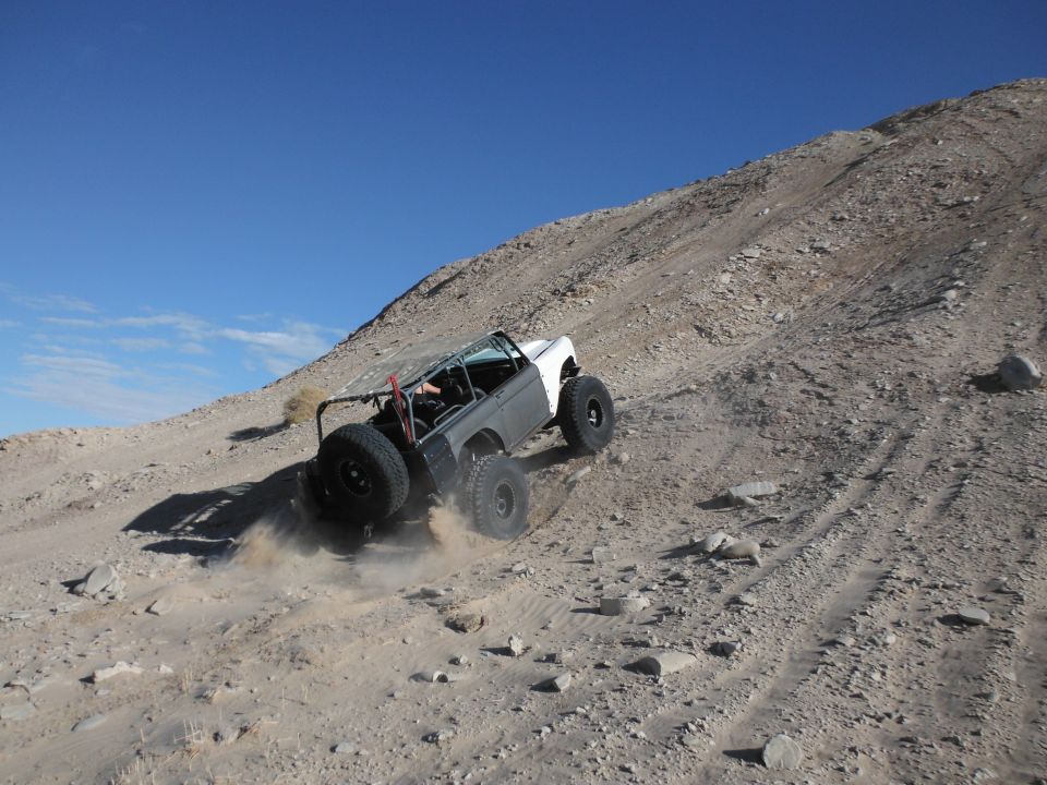 White and grey off-road vehicle with external roll cage and spare tire climbing a dirt hill, kicking up dust