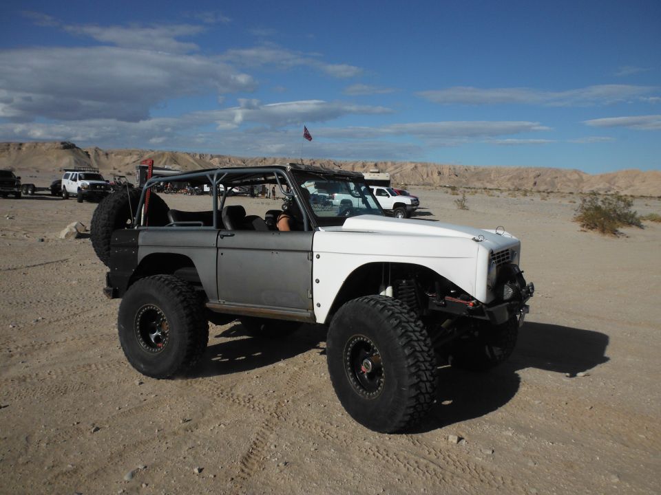 Gray and white modified Ford Bronco with roll cage and large tires parked in a desert landscape