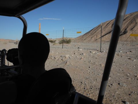 1979 Ford Bronco - View from inside a vehicle as it travels through a dry, rocky desert landscape with a fence line and a distant hill.