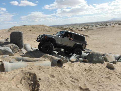 1979 Ford Bronco - A silver Jeep Wrangler drives over concrete pipes and rocks in a sandy desert landscape under a cloudy sky.