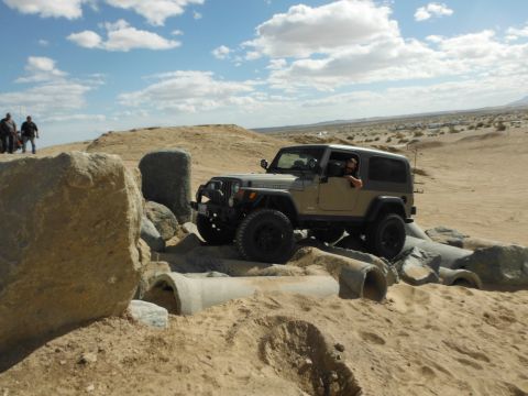 1979 Ford Bronco - A tan Jeep Wrangler tackles a challenging rocky course in a desert landscape under a partly cloudy sky.