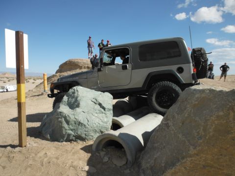 1979 Ford Bronco - A Jeep Wrangler Rubicon is driven over obstacles on a sunny day with spectators watching.