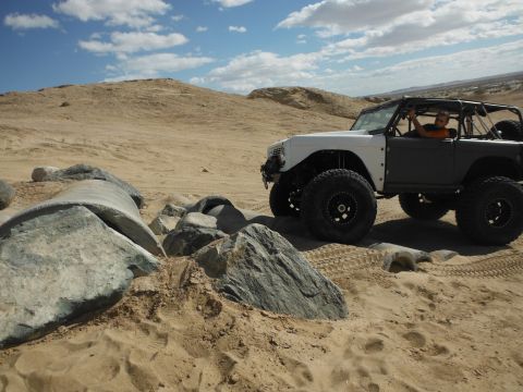1979 Ford Bronco - A heavily modified off-road vehicle navigates a sandy, rocky terrain under a partly cloudy sky.
