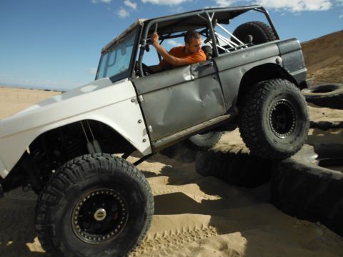 1979 Ford Bronco - A heavily modified off-road vehicle navigates a tire obstacle in a sandy terrain under a clear blue sky.