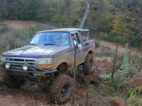 1996 Ford Bronco - This lifted SUV is navigating a challenging off-road trail covered in mud and rocks.