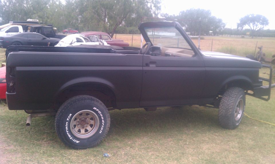 1988 Ford Bronco convertible side view, matte black paint, aftermarket front bumper. Other vehicles visible in background