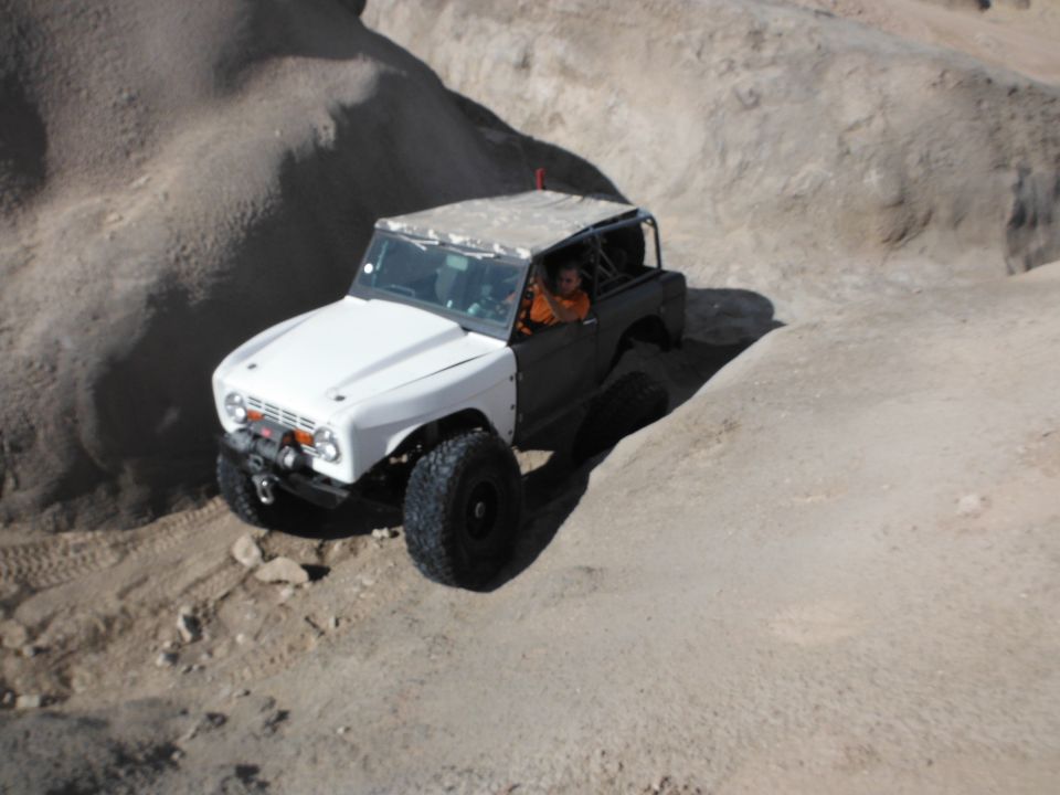 White and dark gray off-road vehicle driving on a dirt trail in a rocky, desert-like area. Driver is visible