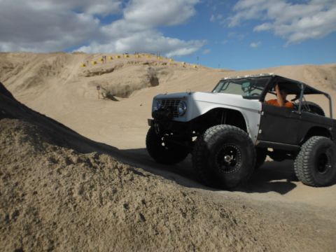 1979 Ford Bronco - Off-road Bronco with custom roll cage navigates a sandy terrain under a cloudy sky.