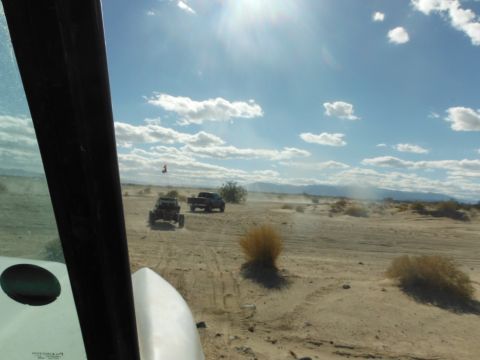 1979 Ford Bronco - Two off-road vehicles traverse a sandy desert terrain, kicking up dust under a bright, partly cloudy sky.