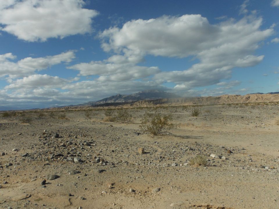 Desert landscape with sparse bushes, rocky ground, distant mountains, and cloudy sky. Watermark reads SUPERMOTORS