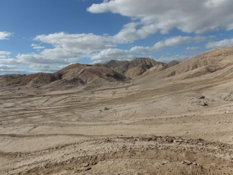 1979 Ford Bronco - A vast, arid landscape stretches towards hills under a partly cloudy sky, marked by numerous tire tracks in the dirt.