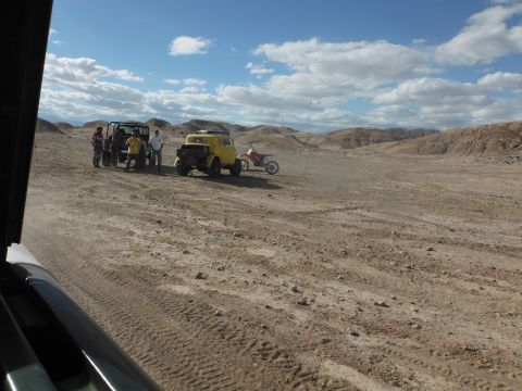 1979 Ford Bronco - Several off-road vehicles, including a yellow vintage truck and dirt bikes, are gathered in a desert landscape under a cloudy sky.