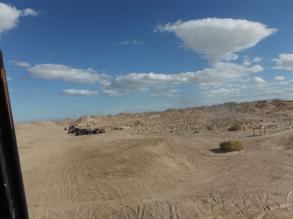 Several off-road vehicles parked in a sandy desert area with hills and a blue sky with clouds