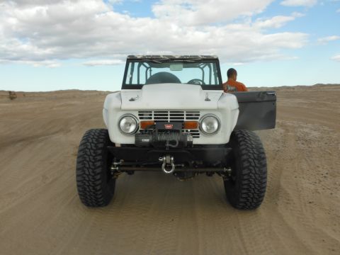 1979 Ford Bronco - A white 1979 Ford Bronco is shown from the front, parked on a sandy terrain under a cloudy sky. The vehicle features large off-road tires,…