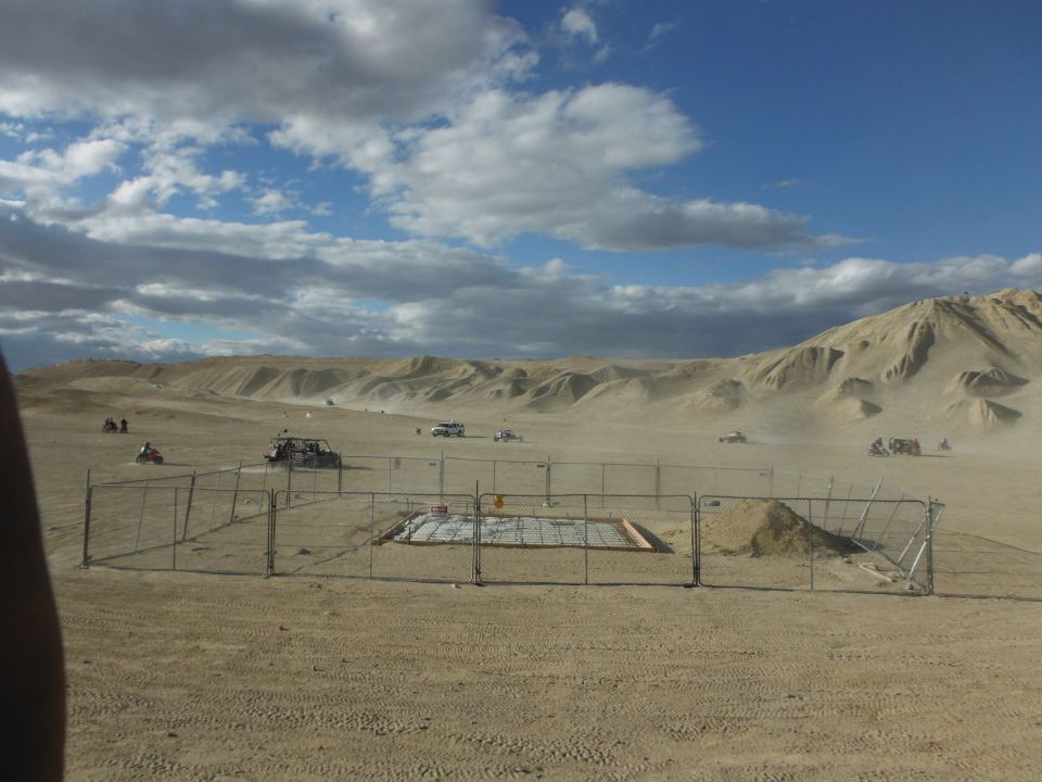 Desert scene with sand dunes, multiple ATVs and off-road vehicles, a fenced enclosure, and a cloudy sky