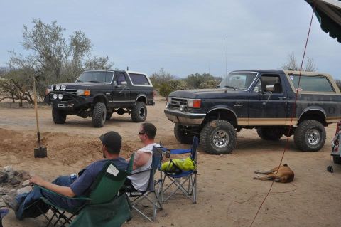 1991 Ford Bronco - Two Ford Broncos are parked in a desert setting, with people relaxing nearby.
