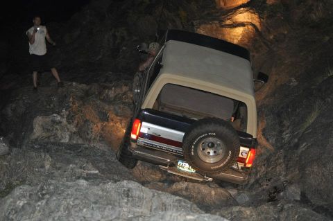 1991 Ford Bronco - A Ford Bronco is shown off-roading at night, with one person observing and another inside the vehicle.