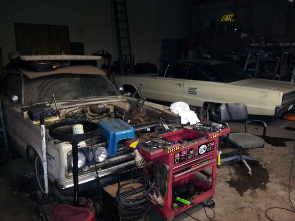 Two cars, one with hood open and engine exposed, in a cluttered garage with a tool cart