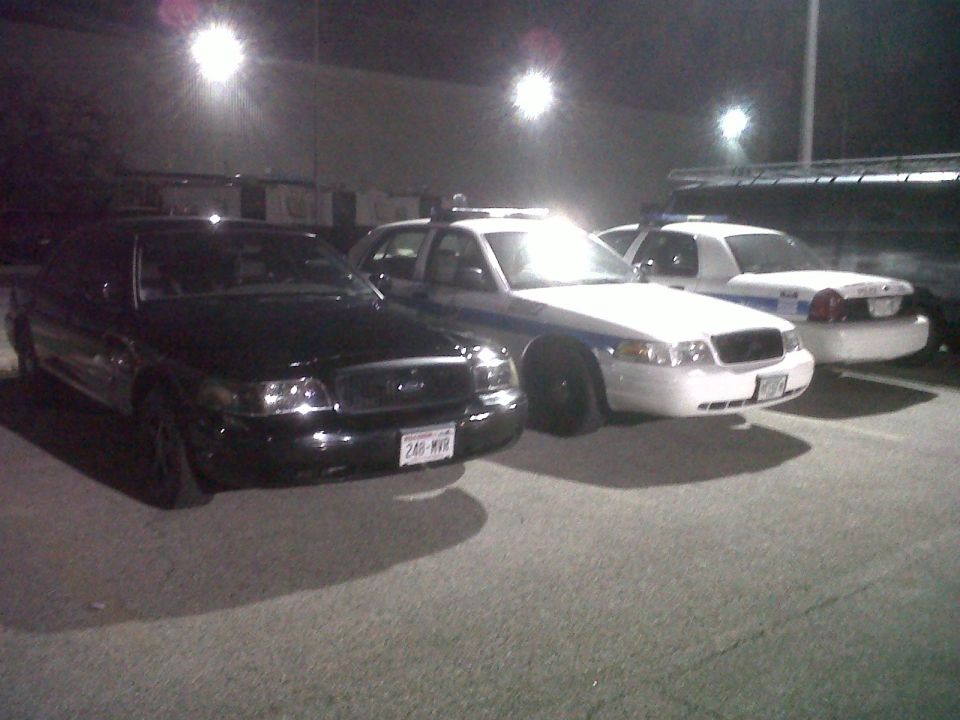 Black Crown Victoria parked beside two white police Crown Victorias in a parking lot at night