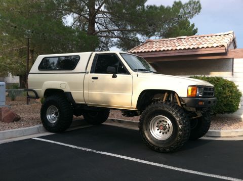 1985 Toyota Pickup/SR5 - This lifted 1985 Toyota Pickup/SR5 features a camper shell and aggressive off-road tires, viewed from the front three-quarter angle.
