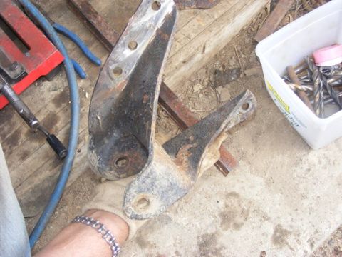 1996 Ford Bronco - Two heavily rusted metal brackets are shown on a concrete surface, alongside various tools and a white container filled with hardware.