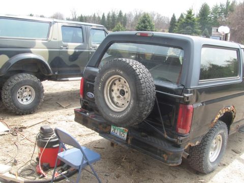 1996 Ford Bronco - Two Ford Broncos are parked outside, one black and one camouflage. The black Bronco shows rust on the rear quarter panel.