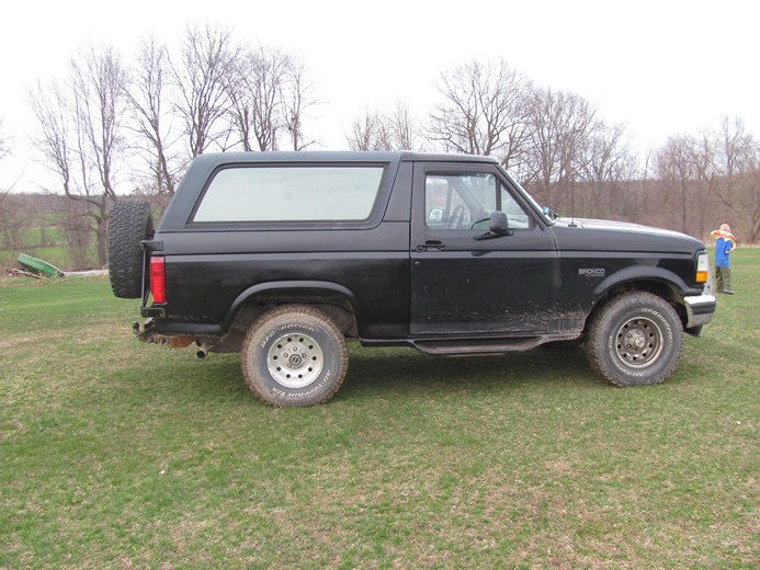 Black Ford Bronco parked on grass, spare tire on back. Someone is standing in the background