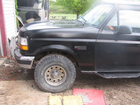 1996 Ford Bronco - The front quarter panel of a black Ford Bronco is visible, with a camouflage vehicle in the background.